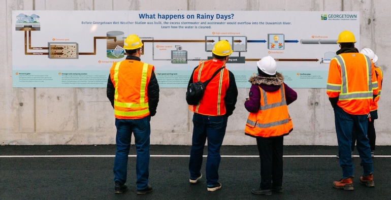 Four people in orange vests and hard hats taking a tour, stop in front of a poster "What happens on rainy days" that explains the treatment process. 