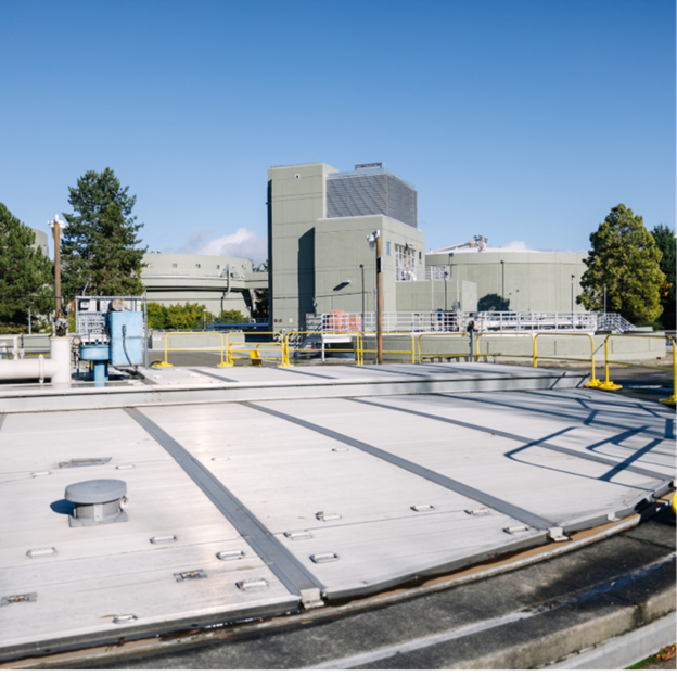 A circular lid to an underground tank is visible in the foreground with a taller narrow concrete building behind it. 