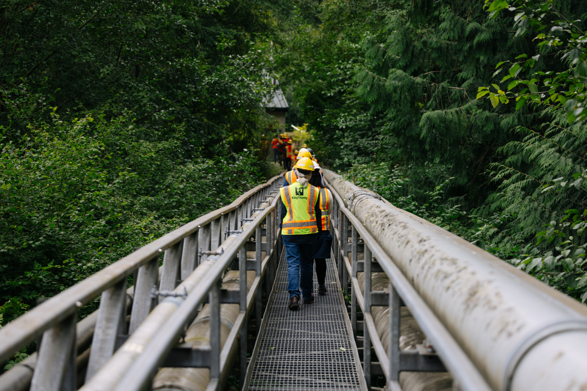 Workers in safety gear walk across a bridge next to a large wastewater pipe surrounded by trees on Vashon Island. The pipe carries wastewater to the Vashon Treatment Plant for cleaning. 
