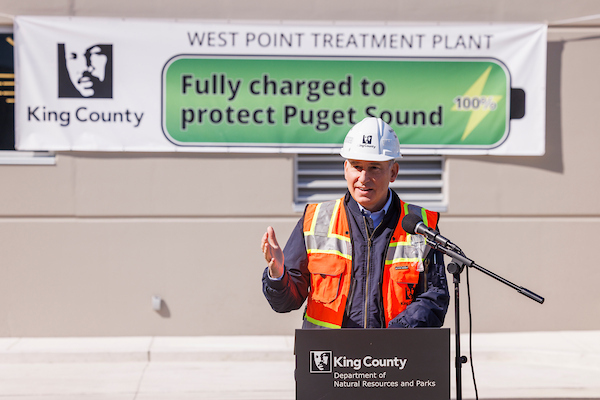King County Executive Dow Constantine, wearing an orange vest and hard hat, at a podium celebrating the completion of the Power Quality Improvement (PQI) project at West Point Treatment Plant.