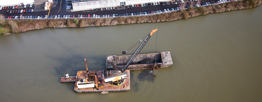 This photo shows two barges in a river. The crane is lifting clean sand out of another barge and placing it in the river to cap sediment contamination to keep it away from animals that live in the river.