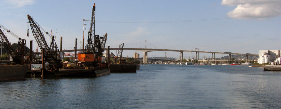 This is a photo showing a cluster of barges on a waterbody with construction equipment on them. The West Seattle Bridge is in the background.