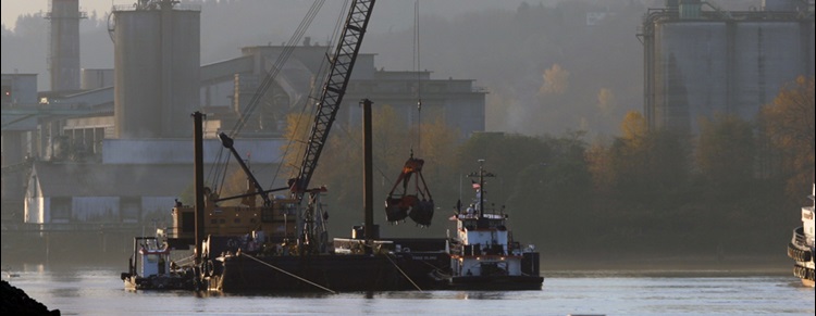 This photo shows a barge with a large mechanical crane on it. The crane is picking up clean sand from another barge and placing it in the river.