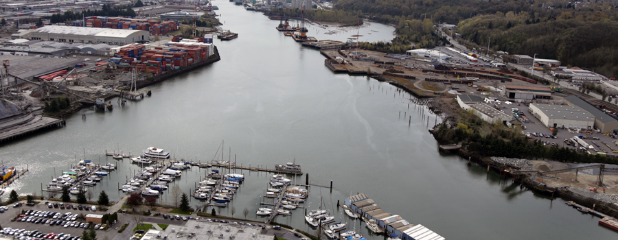 This is a photo of a waterway that splits to the left and right of the photo. In the middle is a marina with sailboats. The left and right banks of the waterway are densely populated with industrial buildings and parking lots. 