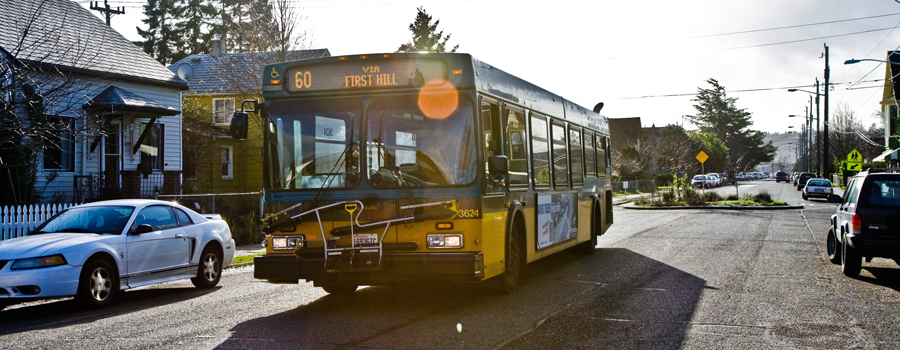 This photo shows the #60 King County Metro bus to First Hill driving down the center of a road in the South Park neighborhood. There are cars parked on both sides of the road.