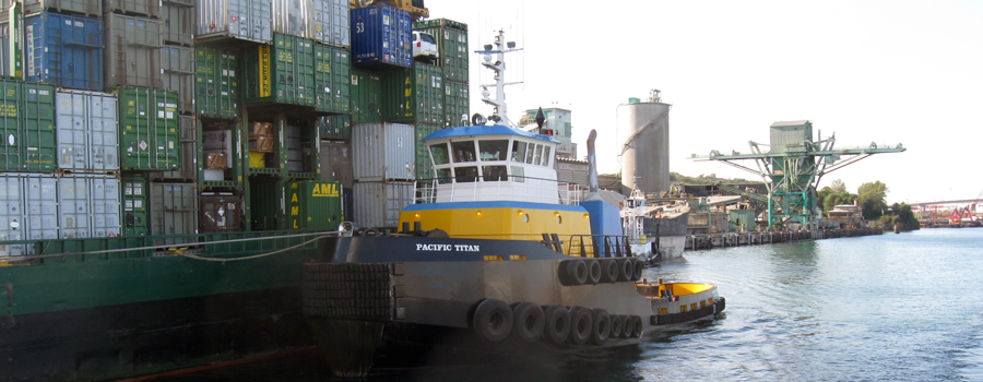 This is a photo showing a part of a barge with multicolored containers on it and in the middle of the photo is a tugboat preparing to move the barge. 