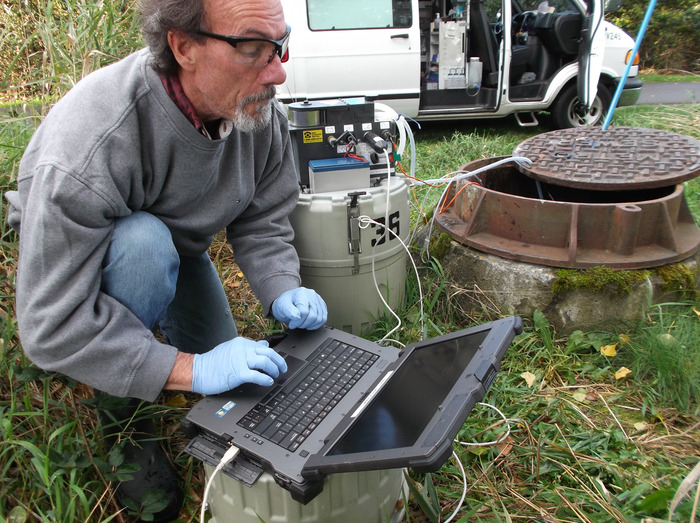 Industrial waste employee sampling a manhole, processing data on laptop nearby wearing latex gloves and protective glasses.