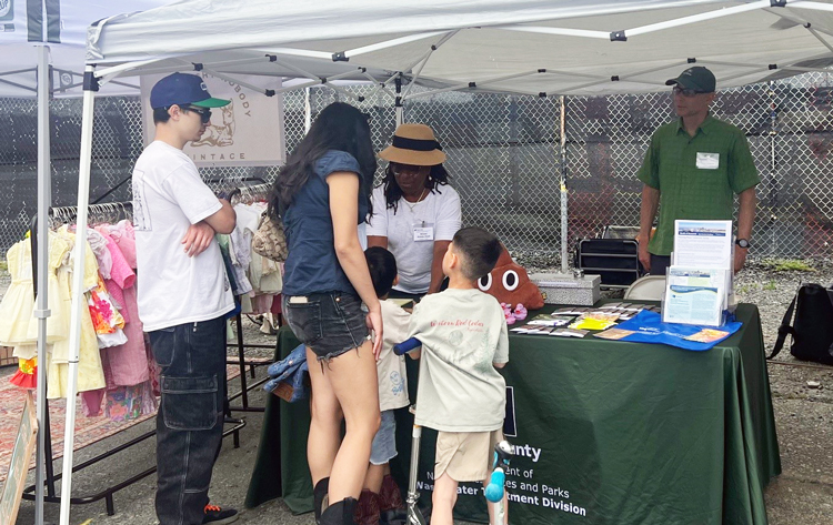 Two adults and two children stand at an information booth under a tent outside while one King County staff members shares project newsletters and giveaways and another watches.