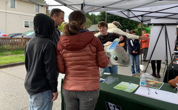 One person in a salmon costume stands behind a table and holds a flyer while talking to three other people under a canopy.