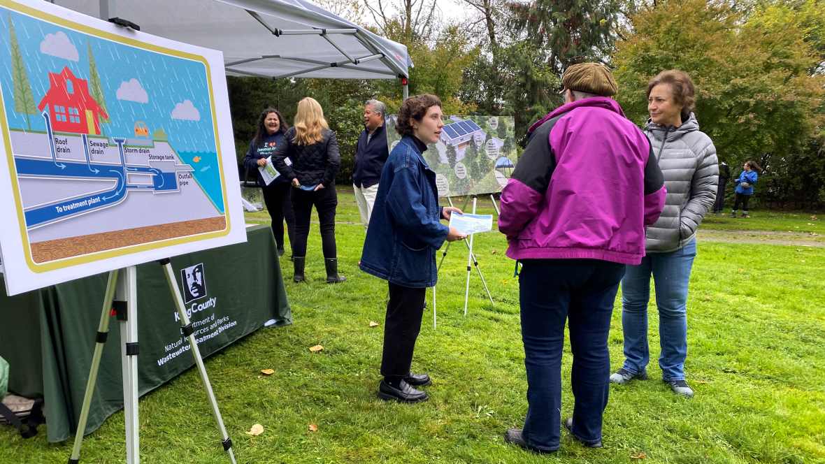 King County staff speak with community members beside educational displays about stormwater and wastewater systems at an outdoor Duwamish Alive event.