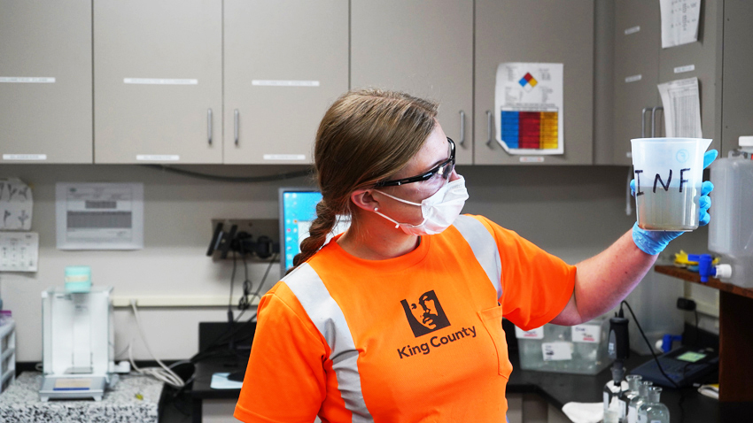Researcher in orange King County official clothing holding up a beaker of biosolids while wearing protective safety glasses and a mask.