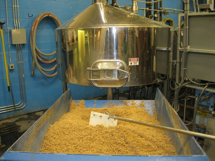 A large container with brewery grain, a rake and industrial tank in the background
