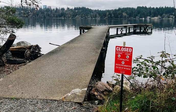 Concrete dock extending into a calm lake on an overcast day, with a red ‘Closed’ sign in the foreground warning that the water is contaminated and to stay out of the water.