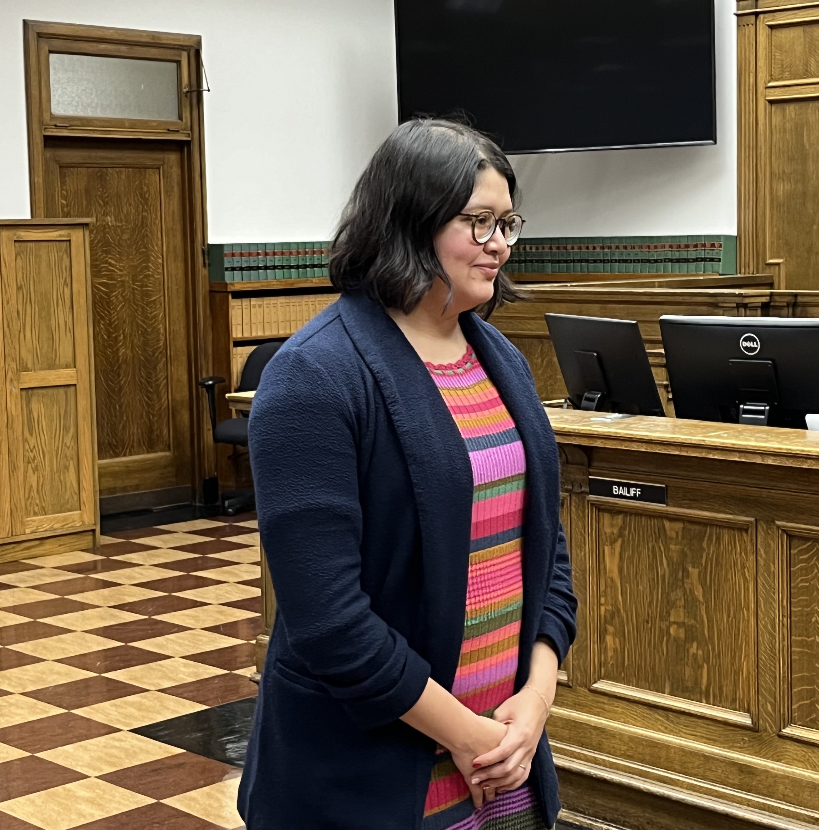 Attorney Selena Alonzo standing in a courtroom with hands clasped in front of her wearing a dark blue jacket and striped shirt 