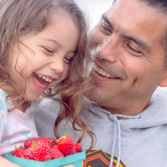 A father holding his young daughter while carrying a carton of strawberries