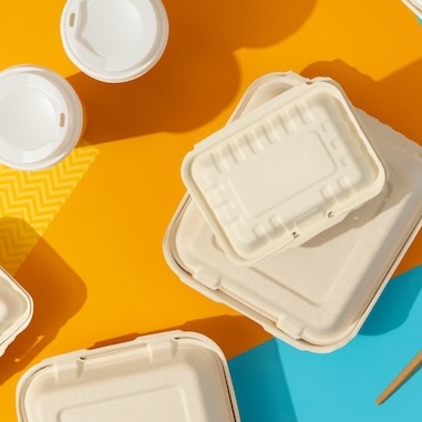 Overhead shot of disposable food containers and utensils on a split yellow and blue background. The containers are made of a light beige material, possibly cardboard or a similar eco-friendly material. There are several containers of varying sizes, some stacked on top of each other. To the left, there are white disposable cup lids. On the blue side, there is a wooden fork and knife, along with a white straw.