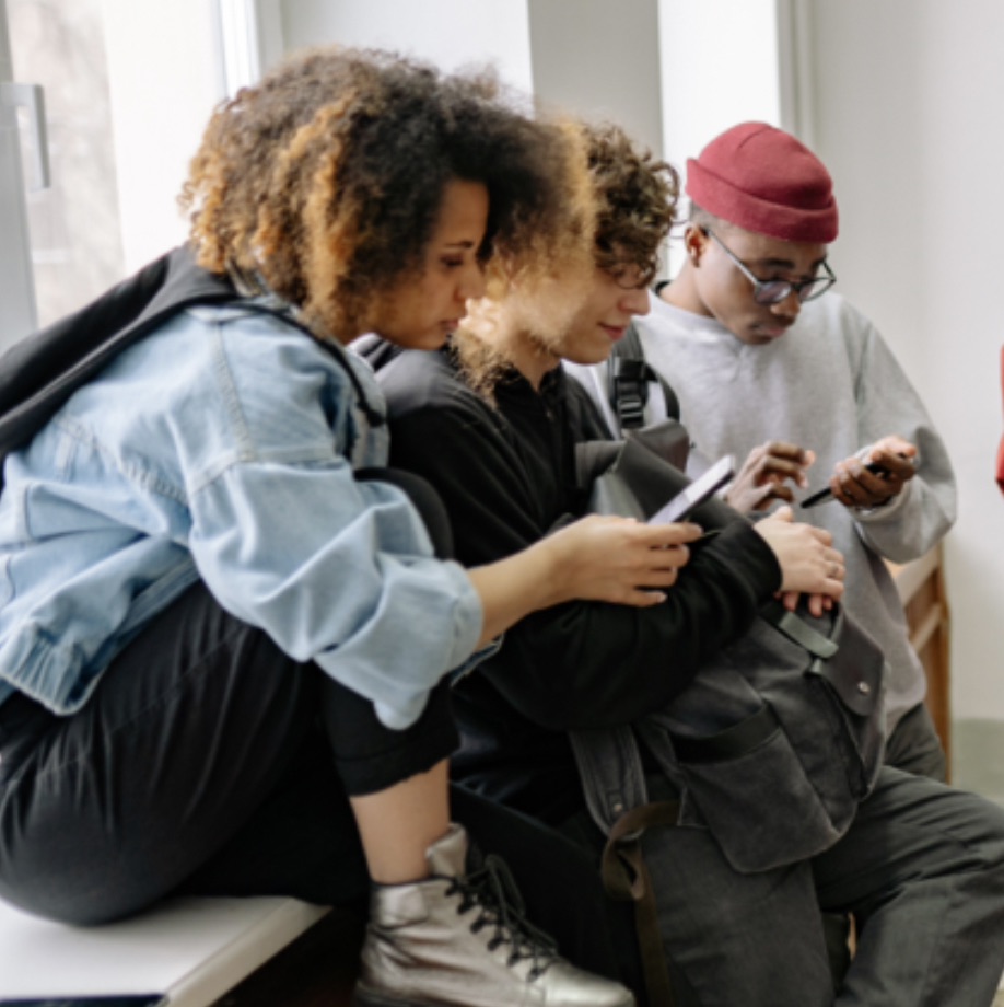 A medium shot of three diverse young adults sitting together indoors near a window, all focused on their smartphones. On the left, a woman with curly hair wears a denim jacket and metallic boots, leaning forward while looking at her phone. In the center, a person with curly hair wears a black hoodie and carries a backpack, also looking down at a phone. On the right, a young man wearing a red beanie, glasses, and a grey sweatshirt looks at his own phone. The lighting is soft and natural, coming from the window behind them, creating a casual, modern atmosphere of friends hanging out.