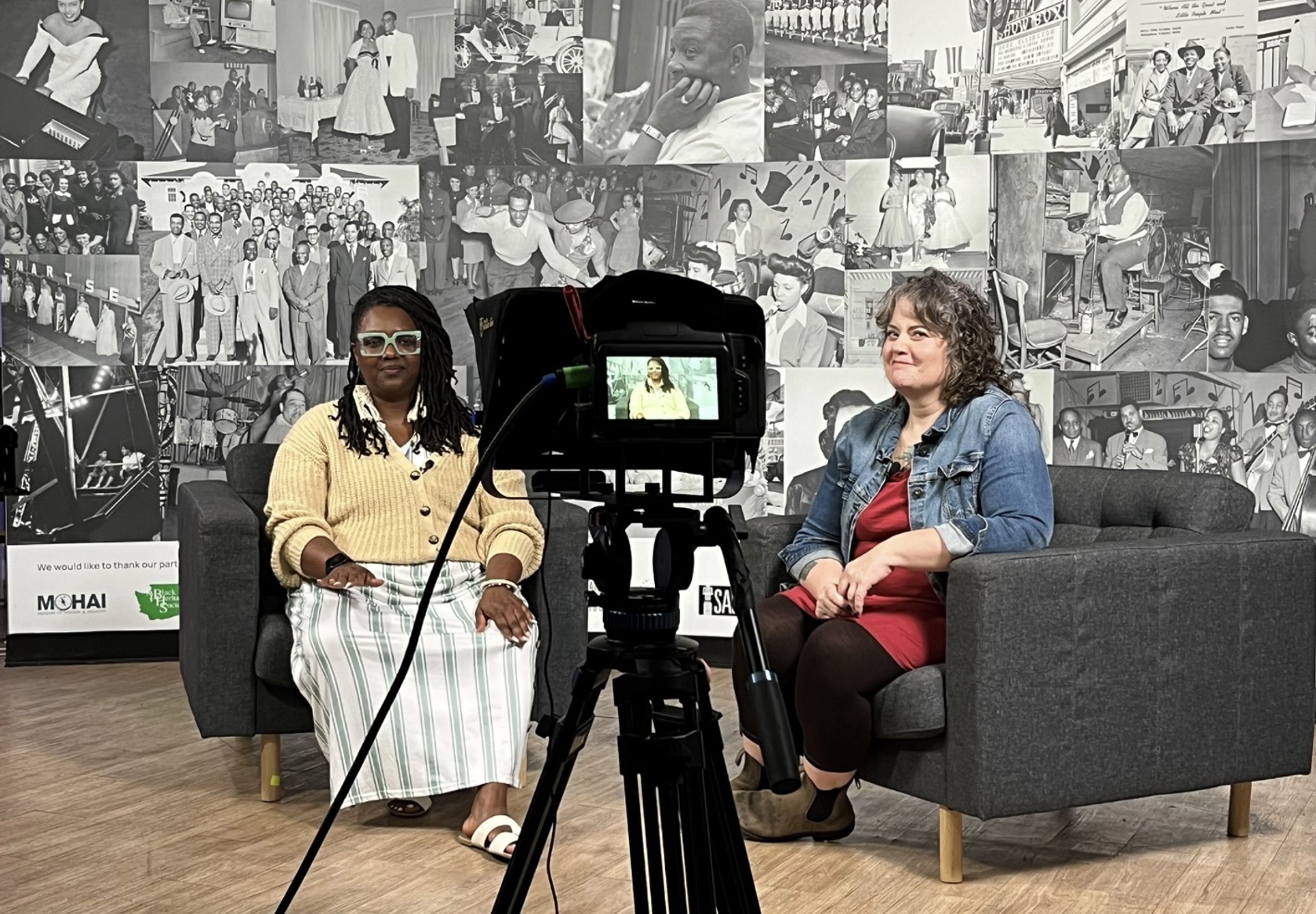 Two women sitting on chairs in front of a professional video camera ready to be interviewed.