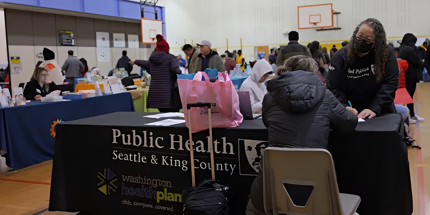 A medium shot a busy indoor health fair, with multiple tables set up and people milling about. In the foreground, a black tablecloth-covered table displays "Public Health Seattle & King County" and "Washington Healthplan." A woman in a black jacket and mask is seated behind this table, appearing to assist someone. 