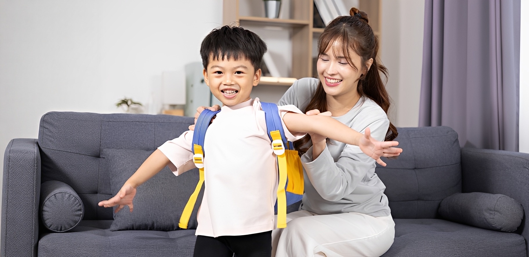 A young mom getting her son ready for school by helping him put his backpack on