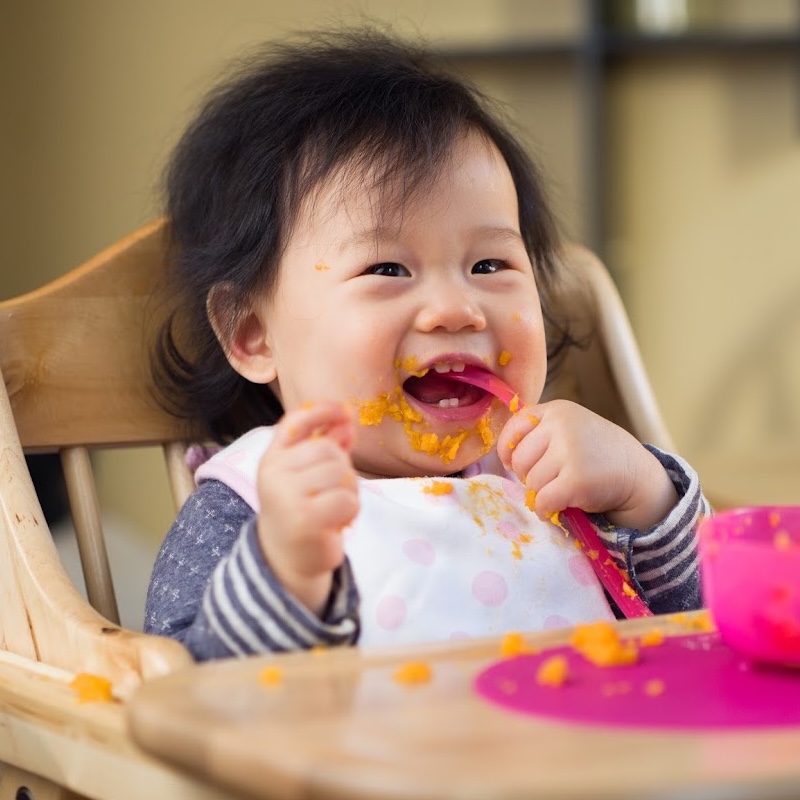 A close-up, eye-level shot shows a happy baby girl sitting in a wooden high chair, eating pureed carrots. The baby has dark, messy hair and is wearing a white bib with pink polka dots and a gray and white striped long-sleeved shirt. Her face is smeared with orange food, and she is holding a pink spoon in her right hand, with some food on it. A pink bowl is visible to the right of the frame, and a pink placemat with scattered food is on the tray in front of her. 