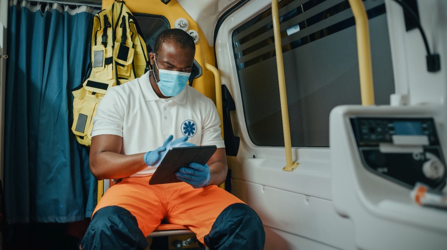 A paramedic wearing a light blue face mask, blue gloves, a white polo shirt with a blue Star of Life emblem, and orange pants sits inside an ambulance. He is looking down and using a tablet computer.