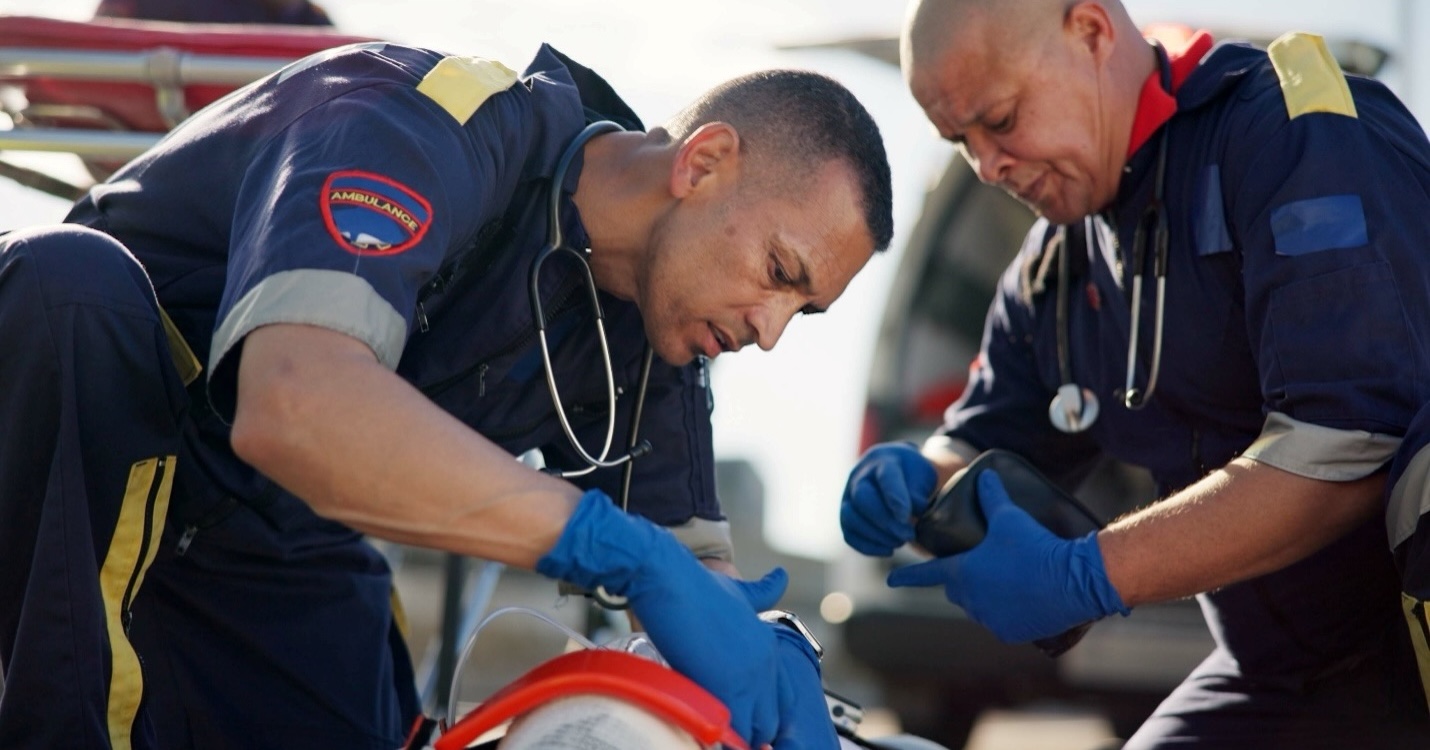 Two paramedics in dark blue uniforms and stethoscopes around their necks are attending to a patient.