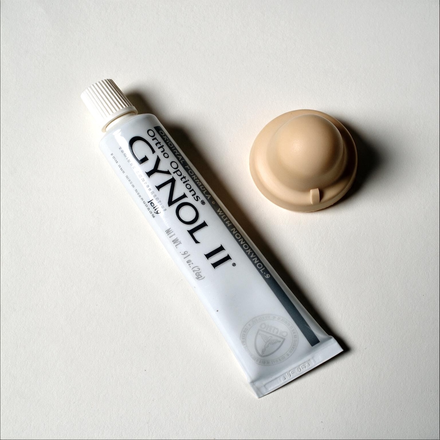 A tube of Gynol II Ortho Options spermicidal jelly and its cap are displayed on a white surface. The tube is oriented diagonally from the bottom left to the top right of the frame, with the cap positioned to the upper right of the tube. The tube is white with black text, and the cap is a light beige color. The lighting is soft and even, creating subtle shadows.