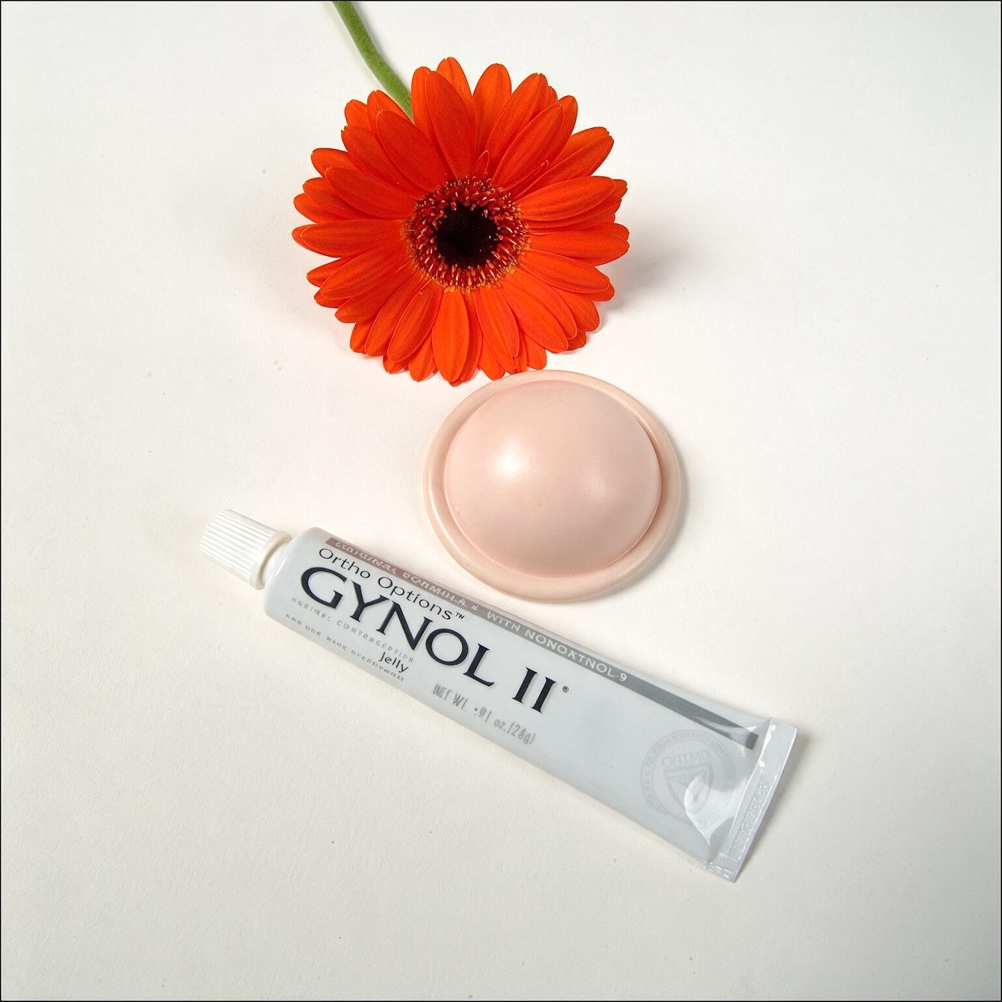 A pale pink diaphragm is placed on a white surface. Below the diaphragm and slightly to the left, a white tube of "GYNOL II" contraceptive jelly is angled diagonally, with its cap pointing towards the top left. 