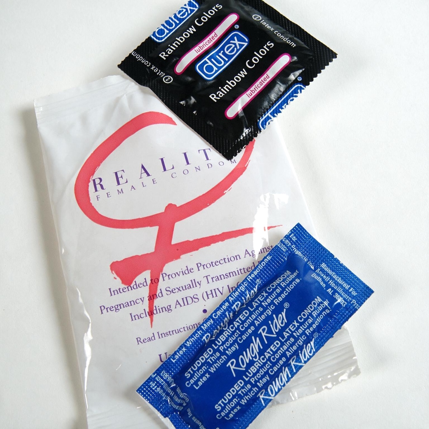 A top-down shot shows a white package of "REALITY FEMALE CONDOM" with a large pink stylized "R" logo. Two black Durex "Rainbow Colors" condom wrappers are placed diagonally on top of the white package, with one slightly overlapping the other. A small blue packet labeled "Rough Rider" is positioned in the lower right corner, partially covering the white package. The background is a plain white surface.
