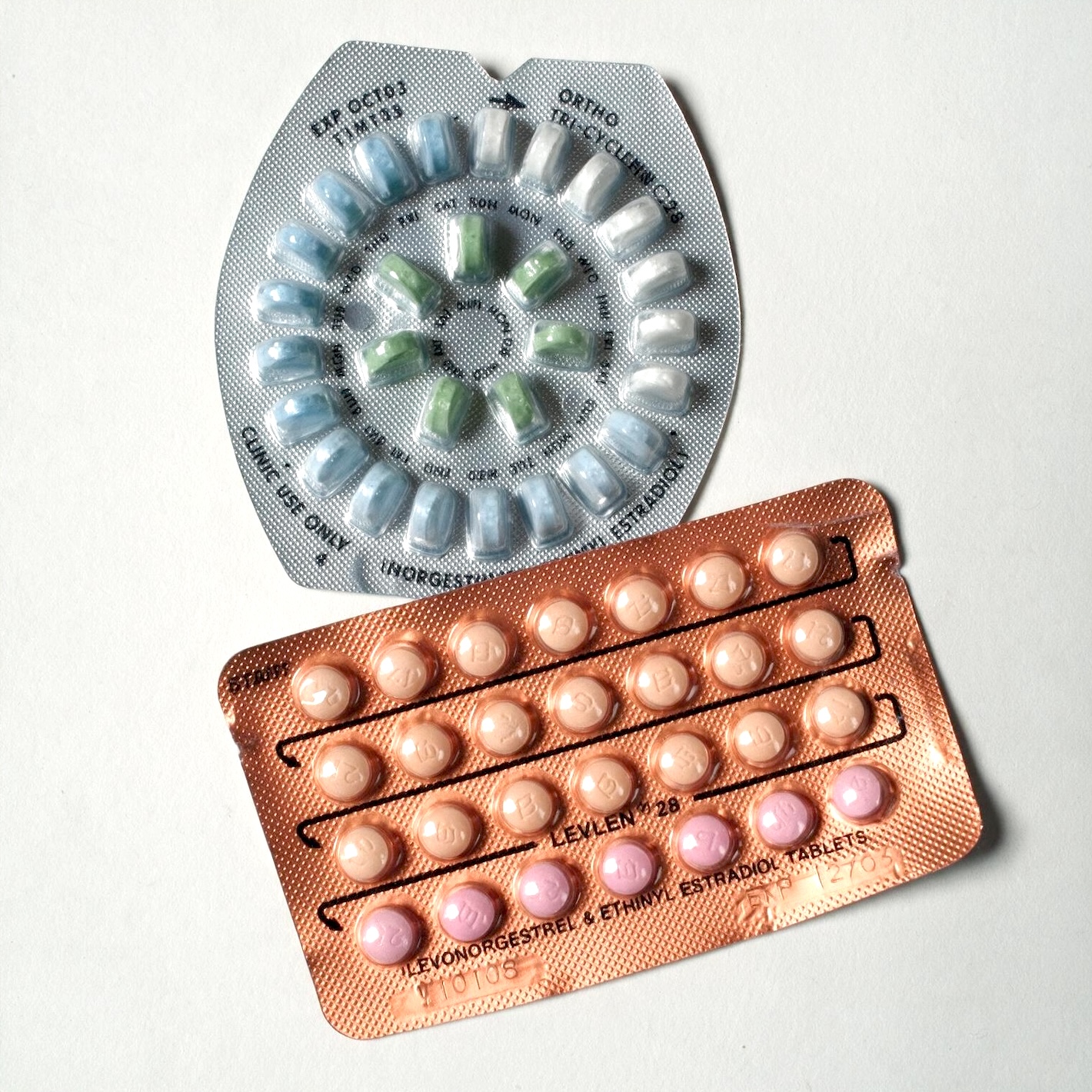 Two blister packs of birth control pills are arranged diagonally on a white surface. The top pack, a silver blister pack, contains 28 pills arranged in a circular pattern. The pills are of three different colors: light blue, green, and white. The pack is labeled "ORTHO TRI-CYCLEN 28" and has text indicating "CLINIC USE ONLY" and "INORGESTREL & ETHINYL ESTRADIOL TABLETS". The bottom pack, a copper-colored blister pack, also contains 28 pills arranged in four rows of seven. These pills are a uniform light peach color, with some appearing slightly pink. 