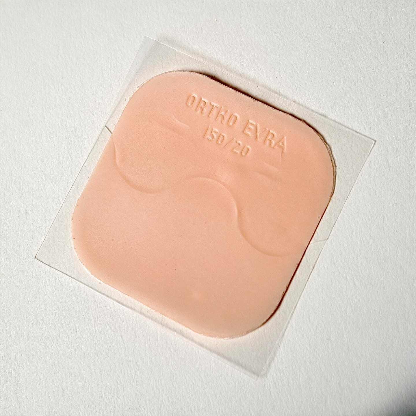A close-up, top-down shot shows a single, light-peach colored contraceptive patch with the words "ORTHO EVRA 150/20" embossed on its surface. The patch is square-shaped with rounded corners and has a slightly wavy, textured surface. It is adhered to a clear, transparent backing, the edges of which extend slightly beyond the patch itself. 