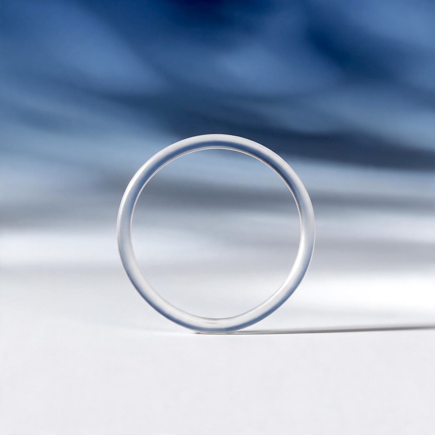 A translucent ring contraceptive is centered on a white surface, casting a soft shadow to its right. The background is a gradient of soft blue, with blurred, wavy patterns suggesting fabric or light. The lighting is soft and diffused, highlighting the translucent quality of the ring.