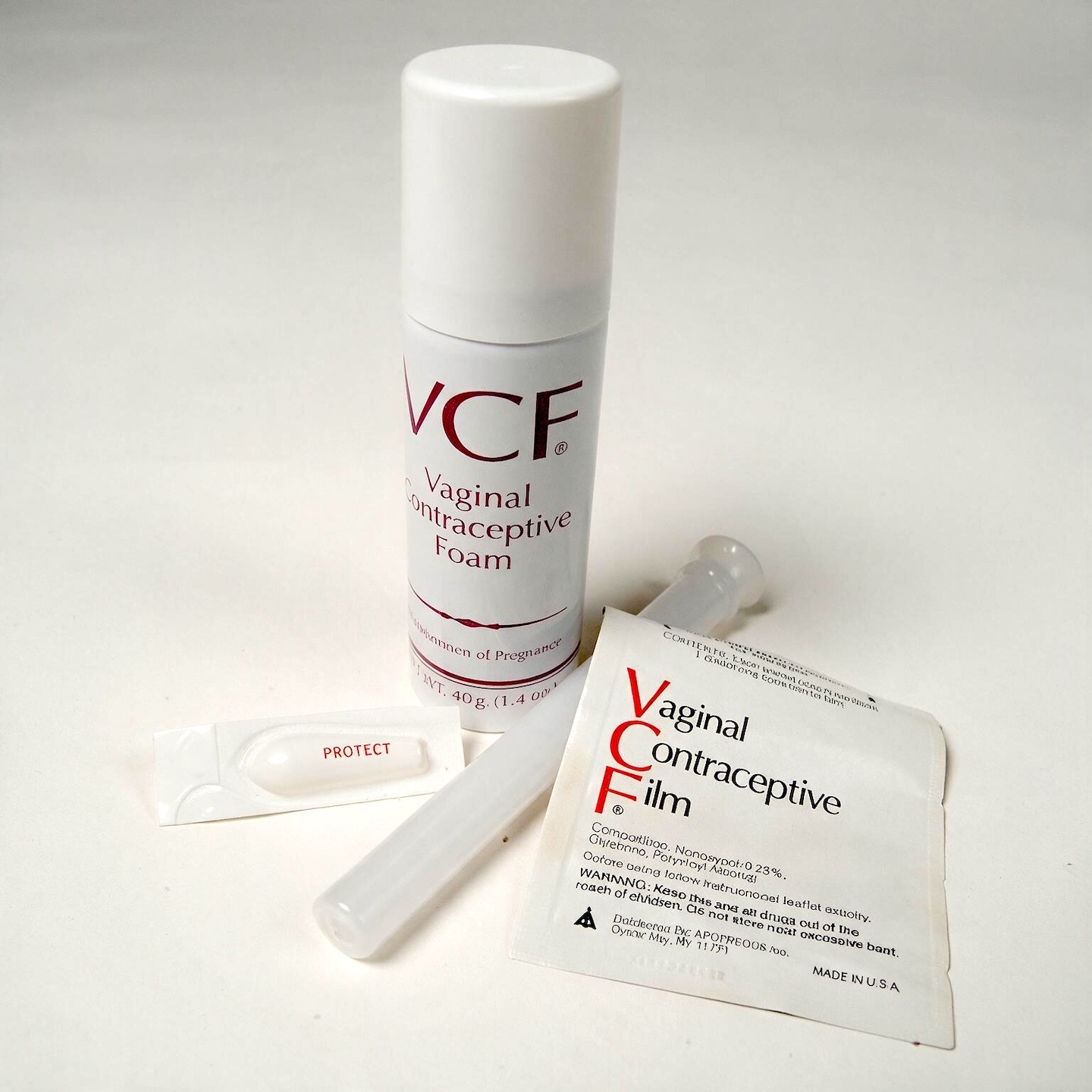 A white, cylindrical can of VCF Vaginal Contraceptive Foam stands upright on a white surface. To the right of the can, a small, white, plastic applicator lies horizontally. To the right of the applicator, a small, white, rectangular packet labeled "VCF Vaginal Contraceptive Film" is also lying horizontally. In the lower left corner, a small, white, individually wrapped applicator with the word "PROTECT" printed on it is visible. 