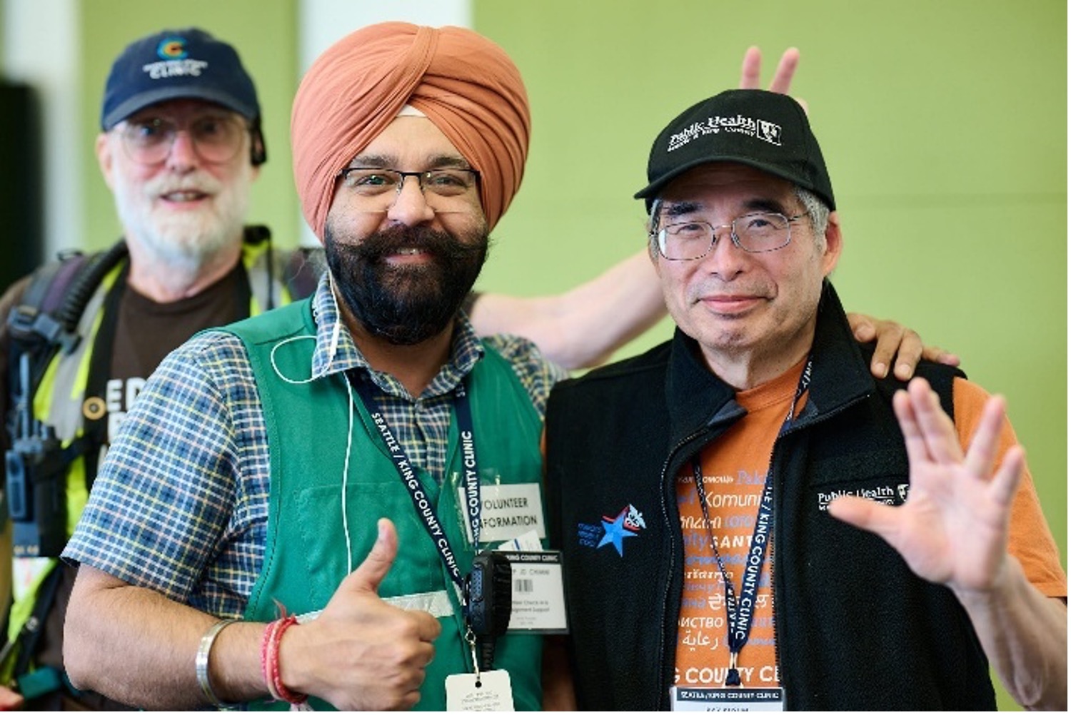 Three men are posing for a photo. The man in the foreground on the left is wearing a blue plaid shirt and a green vest with a "Volunteer Information" badge. He has a turban and a beard and is giving a thumbs-up. The man in the foreground on the right is wearing an orange shirt and a black vest with a "Public Health" logo on his cap. 