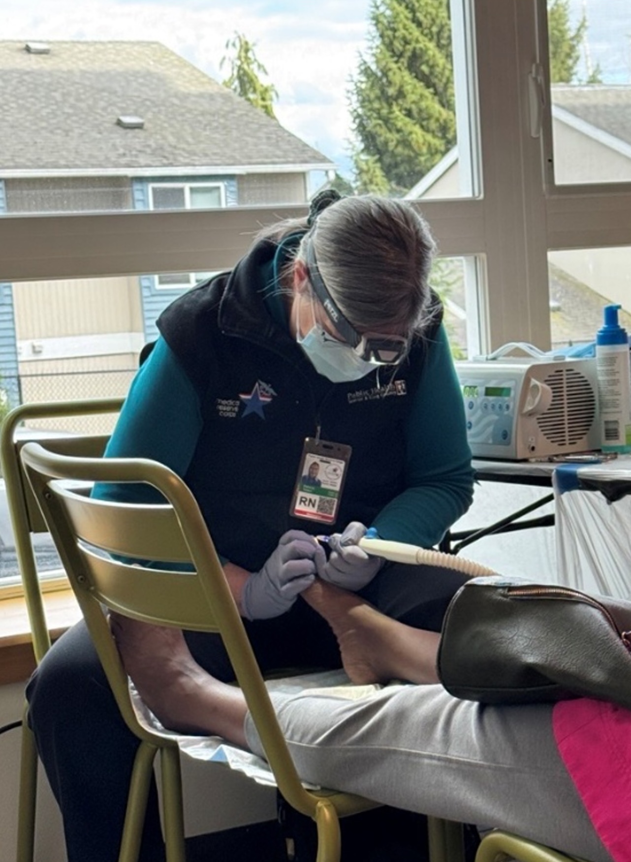 A nurse wearing a mask, goggles, and a vest with an "RN" badge is performing a pedicure on a patient's foot. The nurse is seated on a green chair, and the patient's leg is resting on the nurse's lap. In the background, there is a window with a view of houses and trees, and a table with medical equipment, including a white machine and a bottle of spray. The overall scene appears to be in a clinic or a healthcare setting.