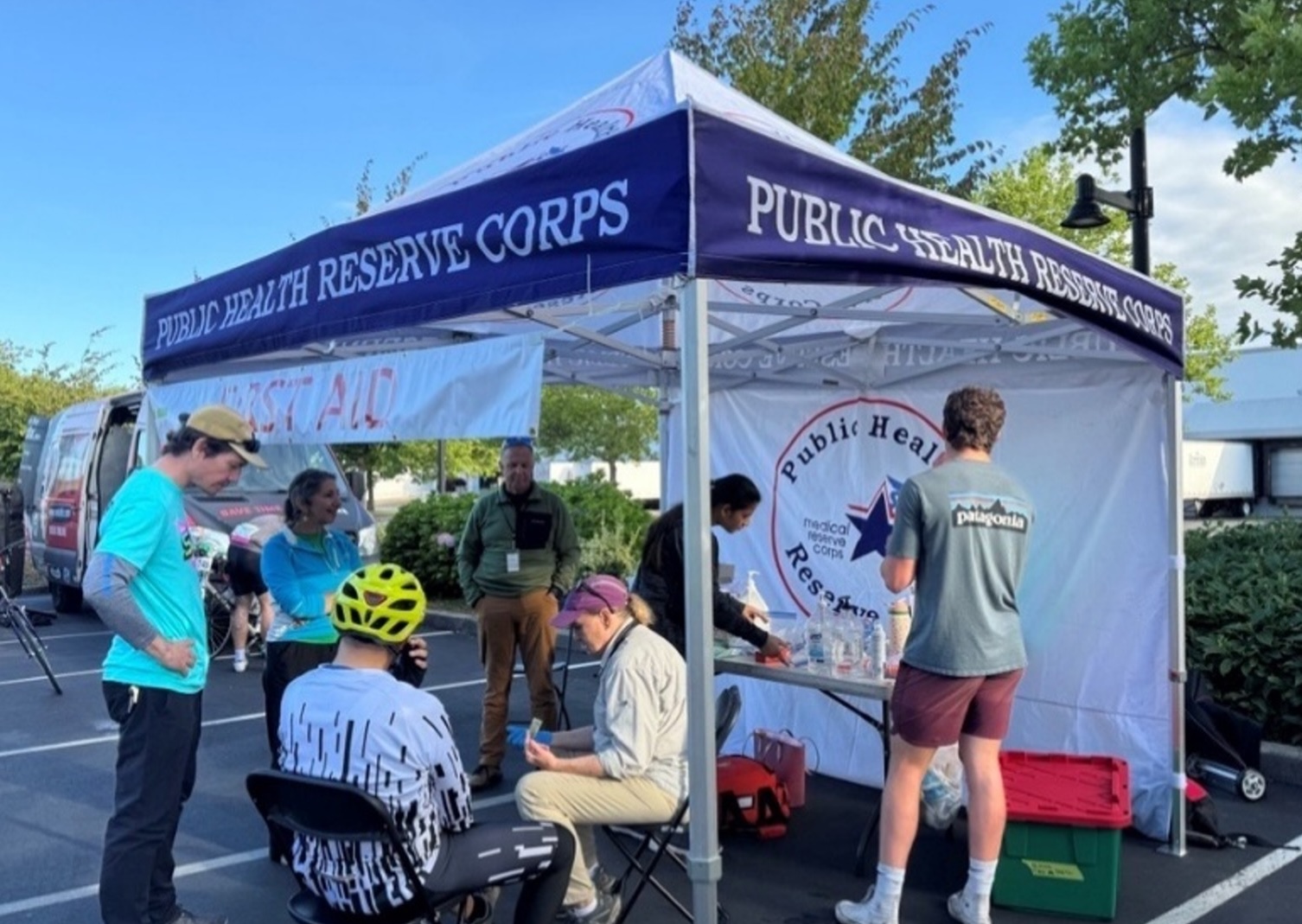A group of people are gathered under a white and blue canopy tent with "PUBLIC HEALTH RESERVE CORPS" printed on it. The tent is set up outdoors, possibly at an event, with a van and trees in the background. Several individuals are interacting, with some appearing to be volunteers or staff. One person is seated and wearing a yellow bicycle helmet. Another person is standing and looking at a table with supplies. The overall scene suggests a public health outreach or first aid station at an outdoor gathering.