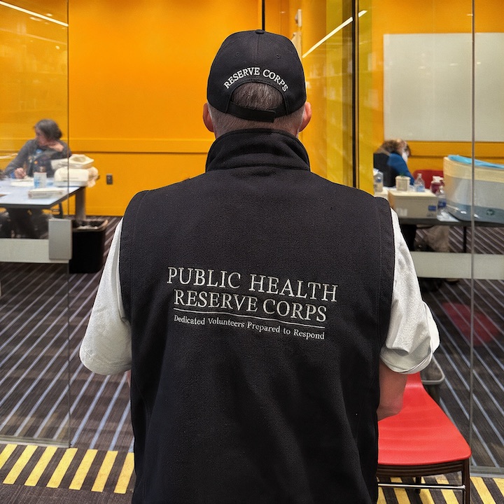 A volunteer is shown wearing a black baseball cap and a black vest with "PUBLIC HEALTH RESERVE CORPS" and "Dedicated Volunteers Prepared to Respond" embroidered on the back.