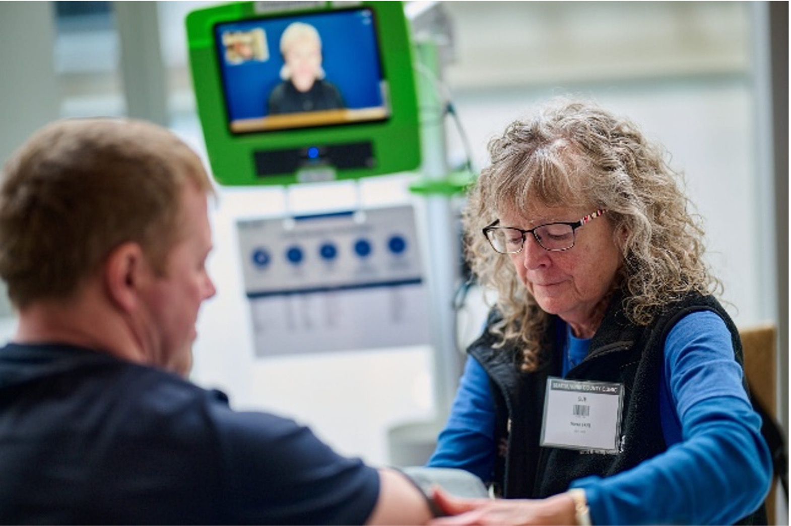 A medium shot captures a healthcare professional, a woman with curly gray hair and glasses, attending to a patient's arm. The woman, wearing a blue shirt and a dark vest with a name tag, is focused on her task. In the background, a man's head and shoulder are visible in profile, facing away from the camera. Above them, a green monitor displays a video call with another person. A medical device with several dials and a chart is also present in the background, suggesting a clinical setting. 