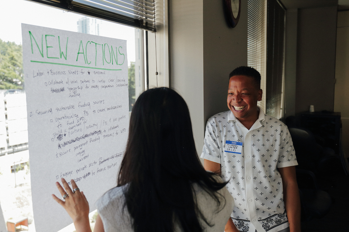 Man smiling while talking, woman writing on paper that says New Actions