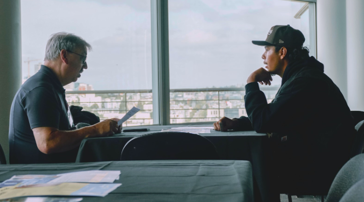 Two men sitting at a table talking. One is holding a stack of papers.