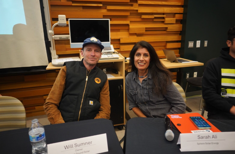A smiling man and woman seated in front of a wooden wall.