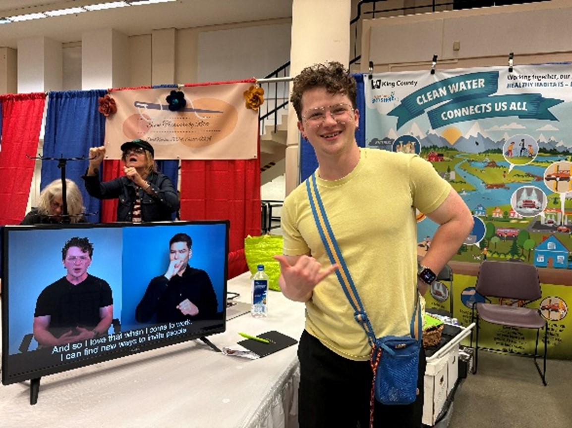 smiling person in yellow shirt next to screen with people talking