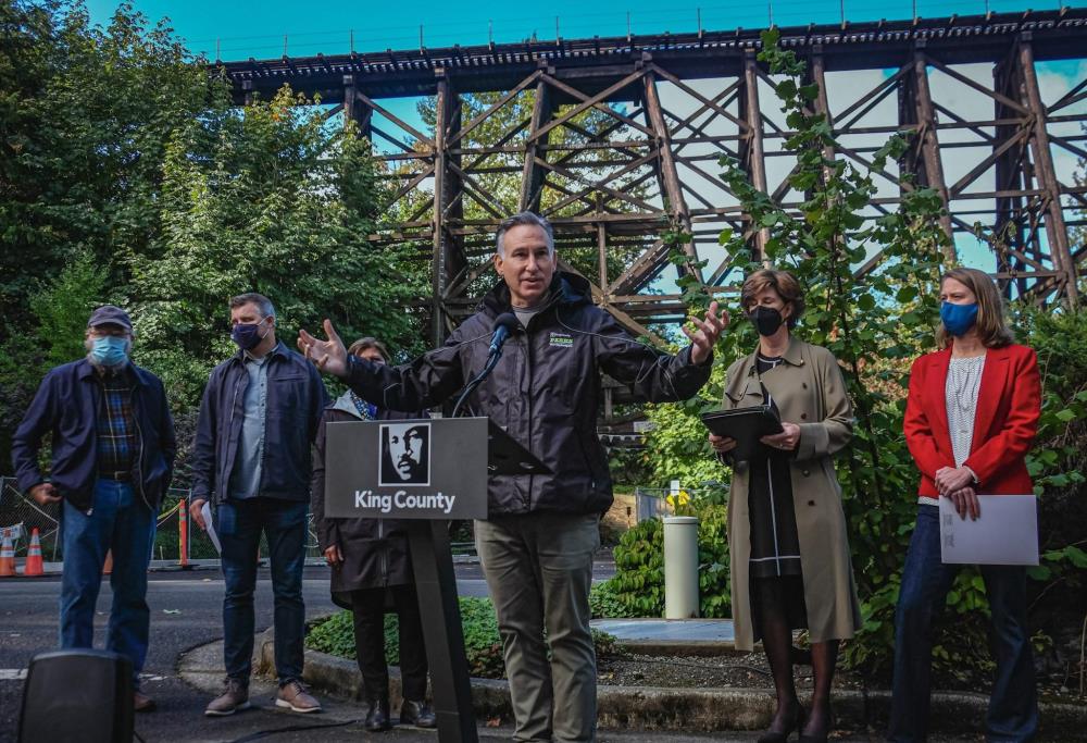 Executive Down Constantine and other leaders speak in front of the Eastrail rail tressle. 