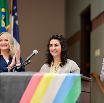 Photo of Executive Shannon Braddock and ADA Specialist, Darya Farivar standing behind a podium with the Disability Pride Flag draped across it. 
