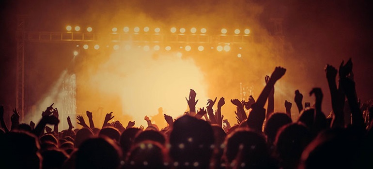 a crowd with their arms raised at a concert. Yellow lights from the stage silhouette the crowd while mist silhouette's the performer.