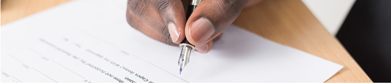 A hand holding a fountain pen over the signature line of a document on a wood table.