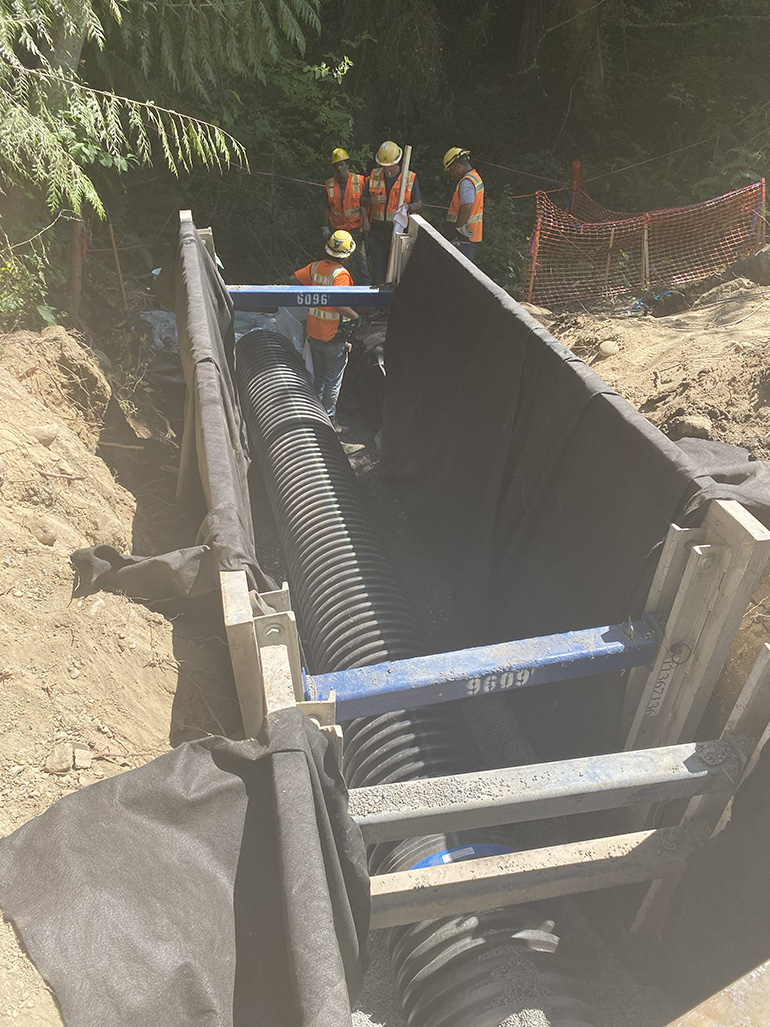 Using a large trench box, four crew members installed the new much larger and longer sturdy thick plastic pipe under the road. 