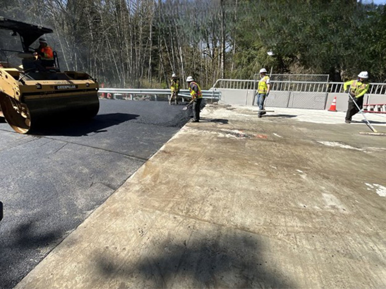 Heavy machinery and crew member standing on newly paved section of the road connects directly to the bridge approach.