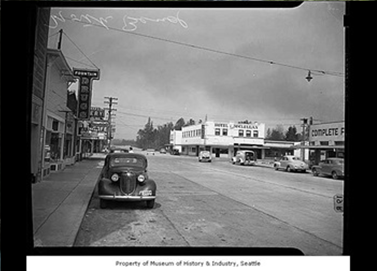 Historical black and white photo with cars parked on the side in front of businesses.
